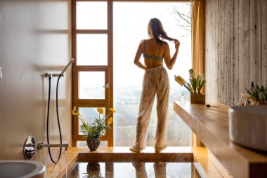 Bir kadın panoramik pencerenin yanında duruyor, nazikçe saçlarını tutuyor. Yumuşak ışık, çiçek açan ahşap dokular sakin bir yansıma ve sabah durgunluğu yaratıyor.