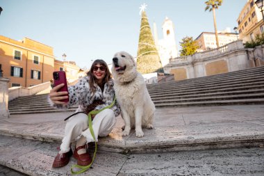 Kadın köpeğiyle birlikte Roma 'daki İspanyol Merdivenleri' nin yanında, arka planda süslü bir Noel ağacı olan şenlikli bir selfie çekiyor. Tatil atmosferi, kış turizmi, neşeli şehir tatili seyahati ve Avrupa