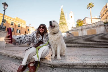 Kadın köpeğiyle birlikte Roma 'daki İspanyol Merdivenleri' nin yanında, arka planda süslü bir Noel ağacı olan şenlikli bir selfie çekiyor. Tatil atmosferi, kış turizmi, neşeli şehir tatili seyahati ve Avrupa
