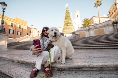Kadın köpeğiyle birlikte Roma 'daki İspanyol Merdivenleri' nin yanında, arka planda süslü bir Noel ağacı olan şenlikli bir selfie çekiyor. Tatil atmosferi, kış turizmi, neşeli şehir tatili seyahati ve Avrupa