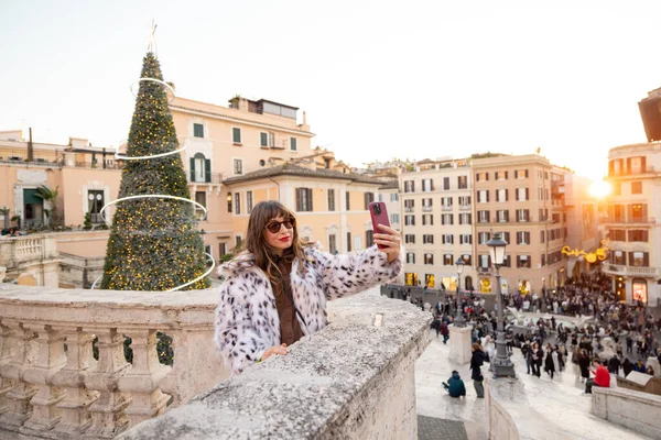 Roma 'ya tepeden bakan, süslü bir Noel ağacı ve canlı bir şehir meydanı olan İspanyol Merdivenleri' nin yanında selfie çeken bir kadın. Tatil atmosferi, kış turizmi, şehir tatili seyahati ve Avrupa