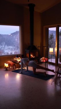 Kışın, sıcacık bir dağ evinde şöminenin yanında yoga yapan bir kadın. Sıcak mum ışığı, sakin atmosfer, yavaş yaşam tarzı, akşam rahatlığı.