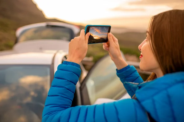 Женщина-путешественница фотографирует восход солнца со смартфоном рядом с машиной — стоковое фото