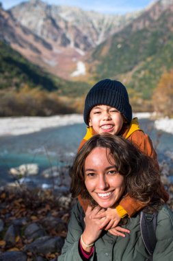 Oğlu, annesi, gezginleri kucaklıyor, sonbahar mevsiminde, güzel bir arazide, nehirler ve dağlarla birlikte, açık havada mutluluk ayakkabıları giyiyor.