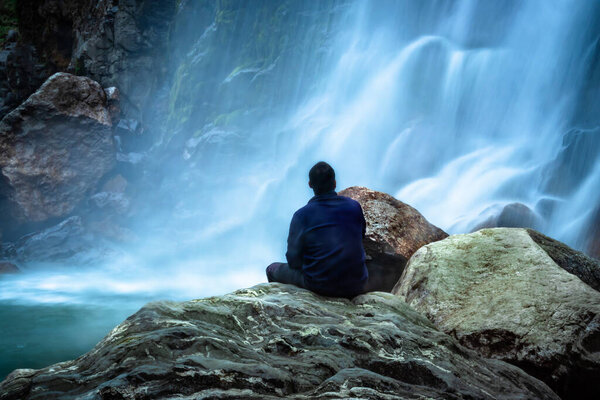 man sitting at rock in front of waterfall white water stream falling from mountains long exposure image is taken at jang waterfall tawang arunachal pradesh.