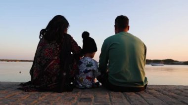 Ebeveynler ve çocuk silueti, alacakaranlıkta suyun yanında dinleniyor.