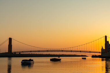 Asma köprü ve deniz kenarındaki siluetlerin yer aldığı resimli günbatımı manzarası