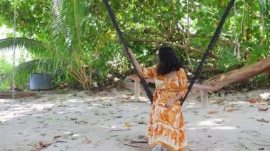 Sahilde arka planda hamakta asılı bir kadının olduğu sakin bir tatil yeri sahnesi.