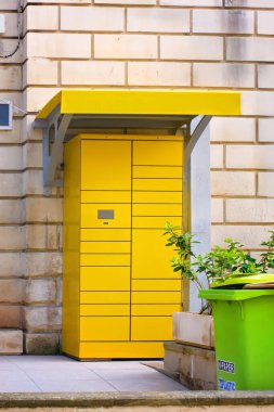 Güneşli bir kentsel alanda, geri dönüşüm kutusu olan bir duvarın yakınındaki modern sarı paket dolabı..