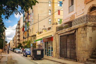 Sliema, Malta - 10.19.2025: Eski ve modern binaların karışımı, yerel bir dükkan, park edilmiş arabalar ve Akdeniz 'de bir geri dönüşüm kutusuyla parlak bir kentsel sokak.