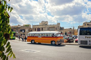 Imdina, Malta - 09.20.2025: Güneşli bir caddede turuncu bir otobüsle otobüs durağı, güzel bulutlu bir gökyüzü altında insanlar ve eski binalar.