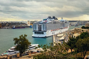 Valletta, Malta - 11.05.2025: MSC Dünya Europa yolcu gemisi ile birlikte MSC Dünya 'nın manzaralı limanının havadan görüntüsü bulutlu bir akşam gökyüzü altında demirlemiş ve etrafı yeşil ağaçlarla çevrili.