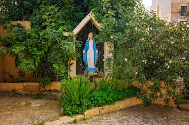 Sliema, Malta - 11.03.2025: Yeşillik ve yol kenarındaki çiçeklerle çevrili Bakire Meryem heykelinin bulunduğu resmi bir Akdeniz avlusu.