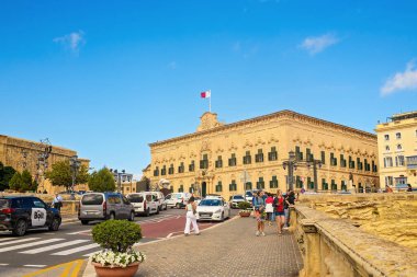 Valletta, Malta - 11.05.2025: Castille Meydanı yakınlarındaki cadde boyunca yürüyen insanlar ve Malta bayrağı altında tarihi bir Barok binası.
