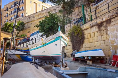 Taştan duvarlar, yeşil santraller, şehir binaları ve parlak güneş ışığıyla çevrili bir kıyı sahasında geleneksel Akdeniz tekneleri..