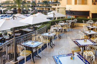 San Giljan, Malta - 11,11.2025: Akdeniz 'de bir şehrin mimarisine karşı dizilmiş beyaz dövmeli demir sandalyeler ve desenli mavi ve sarı masaların yer aldığı manzaralı bir açık kafe terası.