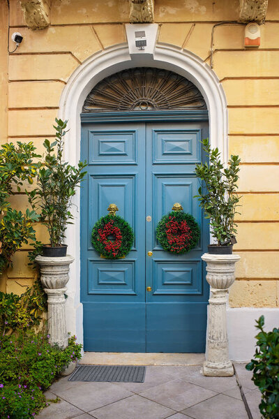 Christmas wreaths made of green fir branches and red berry clusters hang on a blue entrance door in a cozy Maltese courtyard.