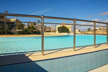 Açık mavi gökyüzünün altında turkuaz su bulunan açık hava yüzme havuzunun krom parmaklıklarından manzaralı bir manzara, kıyı mimarisinin bir zeminine karşı kurulmuş..