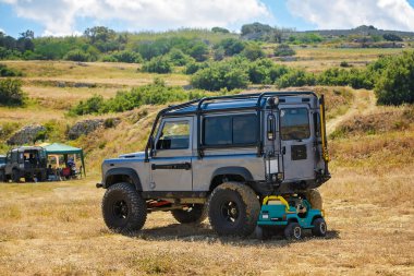 Saint Pauls Körfezi, Malta - 27 Nisan 2025: Modifiye edilmiş gümüş 4x4 Land Rover Defender 'ın yan görüntüsü kırsal kesimdeki bir kamp alanında bir çocuk oyuncak arabasının yanına park edilmiş..