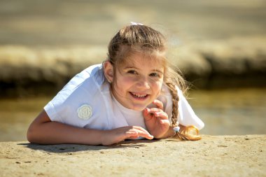 Deniz kenarındaki taşlı bir sahilde uzanmış neşeli bir kızın portresi. Güneşli bir yaz tatilinde kameraya bakıp neşeyle gülümsüyor..