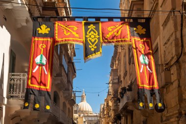 Decorative festive banner with bishop symbols hanging between historic limestone buildings in Valletta, with the iconic church dome in the background during Maltese Festa season.