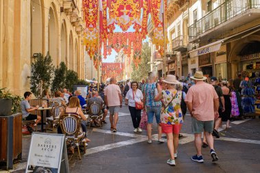 Valletta, Malta - September 17, 2025: View of a busy pedestrian street with tourists and outdoor cafes, decorated with large red and yellow festive banners during the local festa season