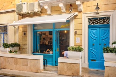 Sliema, Malta - 3 Kasım 2025: İnsanlar, şehir caddesindeki sarı kireçtaşı duvarlara karşı parlak mavi cepheli, rahat bir yerel kafede dinleniyor..