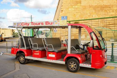 Valletta, Malta - 29 Kasım 2025: Caddeye park etmiş eski bir taş duvarın yanındaki boş, küçük, açık hava elektrik şehir turu otobüsünün yan görüntüsü.