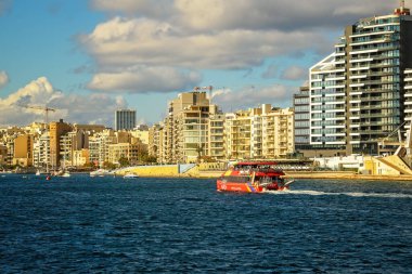 Sliema, Malta - 29 Kasım 2025: Kırmızı bir yolcu feribotu Akdeniz kıyısı boyunca modern mimarisi olan şehir limanını geçiyor.