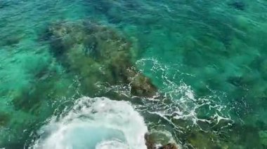 Kristal berraklığında turkuaz deniz suyunun üst görüntüsü. Beyaz köpük dalgaları kıyıya yakın engebeli kayalık bir deniz tabanına çarpıyor..