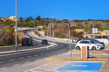 Malta 'daki Ghadira Körfezi plajı yakınlarında viraj otoyolu. Pürüzsüz asfalt yol, arabalar, yol işaretleri, ve engelli park alanı bitkilerin ve mavi gökyüzünün karşısında..