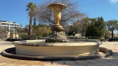 Malta 'daki bir halk parkında melek heykelleri ve büyük dekoratif vazo bulunan antik taş çeşme. Mavi gökyüzünün altında palmiyeler ve yeşilliklerle çevrili zarif mimari anıt..