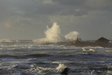 Kıyıdaki fırtına kayaların ve kayalıkların üzerinden büyük bir dalganın sıçrama ve püskürme gördüğünü gösteriyor.