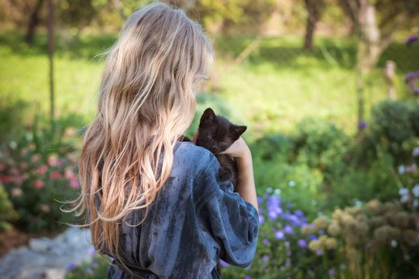 一个金发碧眼的小女孩 肩上抱着一只黑猫 在阳光灿烂的日子里 在五彩缤纷的花园里回头看 — 图库照片