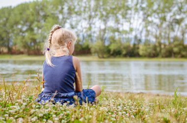 Küçük kız yeşil çayırlarda oturup gölete bakıyor.