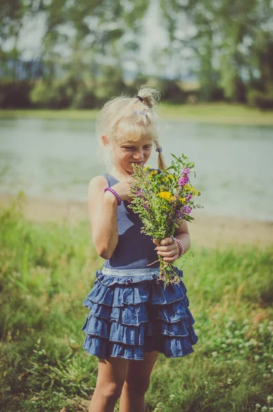 緑の夏の草原の池に花の花束を持つ小さなブロンドの女の子 ストック写真