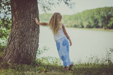 Göletin yanındaki ağacın yanında duran uzun sarı saçlı sevimli çocuk.