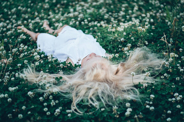 Girl with long blond hair lying in green clover grass with tiny white flowers, view from birds perspective