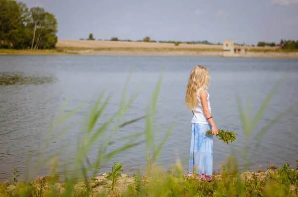 緑の夏の草原の池に花の花束を持つ小さなブロンドの女の子 — ストック写真