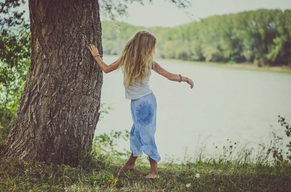 池のそばの木のそばに長いブロンドの髪の子供が立っていて — ストック写真