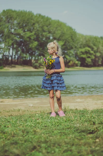 緑の夏の草原の池に花の花束を持つ小さなブロンドの女の子 ストック写真