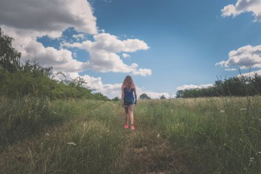 Kırsal yolda yeşil çayırların arasında yürüyen uzun sarı saçlı kız.