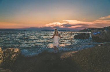 Gün batımında kumsalda tek başına duran, uzun beyaz elbiseli, sevimli bir kız.