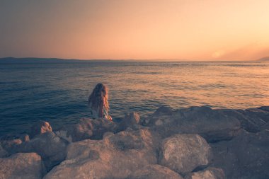 Sevimli kız oturmuş deniz kenarında gün batımını seyrediyor.