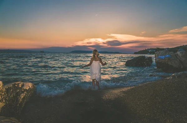 長い白いドレスを着た可愛い女の子一人でビーチに立つ夕日の時青いトーン効果 — ストック写真