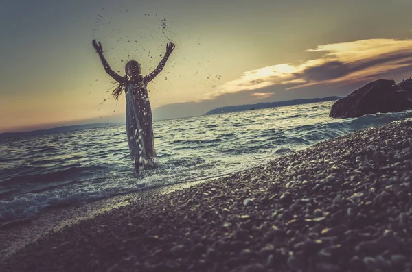 夕日の時間にビーチで海の水で踊る長い白いドレスのかわいい女の子オレンジブルートーン効果 — ストック写真