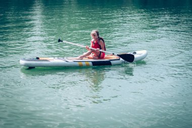 Kırmızı güvenlikli, şişme yelek giyen genç kız yeşil doğada, güneşli bir yaz gününde su yüzeyinde kürek çekiyor.