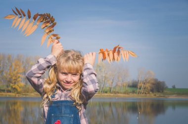 Kot pantolonlu sevimli genç kız. Mavi nehir arka planında parlak turuncu sonbahar yaprakları tutuyor.