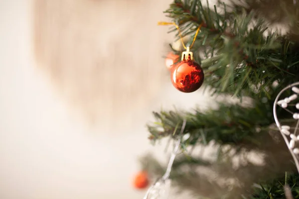 Red round Christmas tree ornament on green branch with copy space and string lights on the background. WInter holidays greeting card. Decorated Christmas tree close up