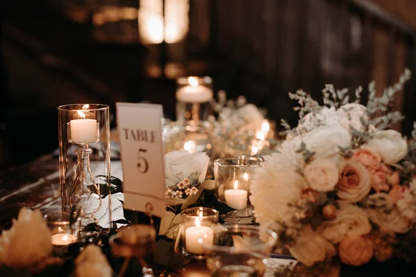 Beautiful luxury rustic wedding guest table decor - wooden tables with glass jars and candles, white roses and eucayptus leaves center pieces on dark background. Luxury loft wedding.